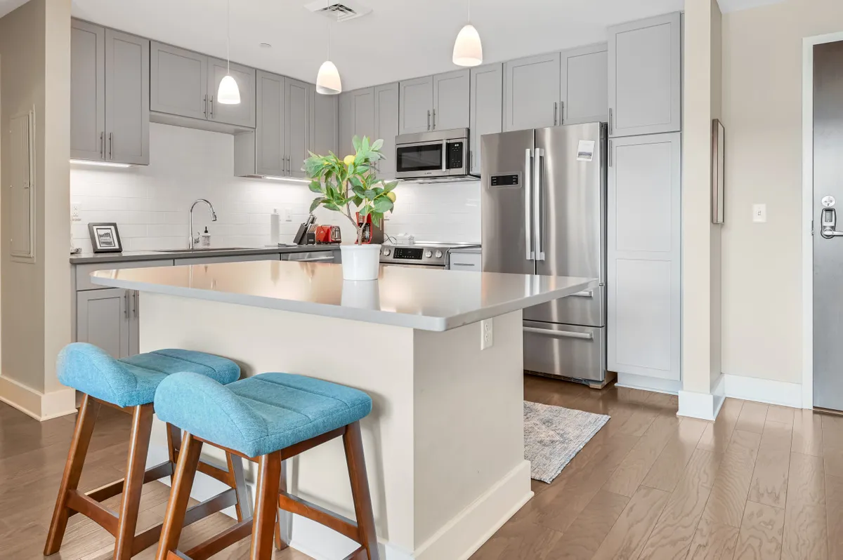 The image features a functional, inviting kitchen space with a prominent island countertop, bar stools for casual seating, modern appliances, and a potted plant adding warmth. The setup encourages both cooking and socializing, reflecting a balance between function and comfort.