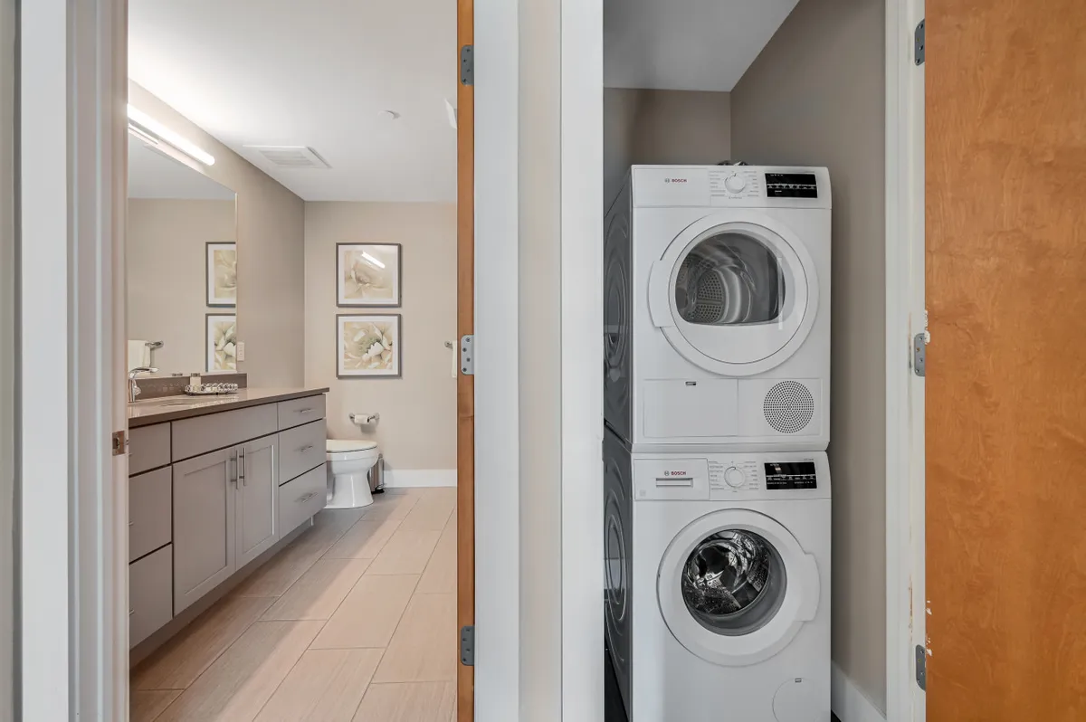 A practical, serene corner of the condo featuring a stacked washer and dryer, a clean bathroom vanity, and framed artwork. The overall mood is calm and organized, highlighting facilities that support daily routines and convenience.