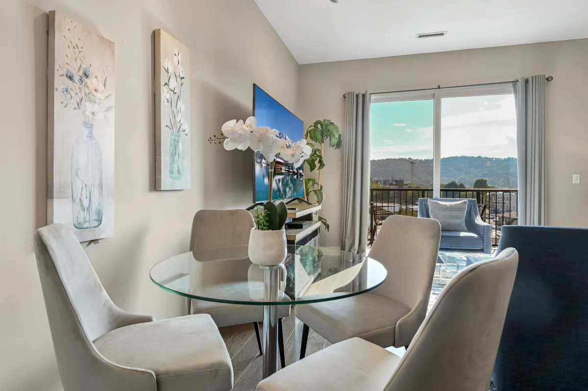 This scene centers around a glass dining table paired with a blue armchair and set near a large window, allowing abundant natural light and views outside. Decorative paintings add a modern touch, and the area is perfect for dining, socializing, or relaxing.