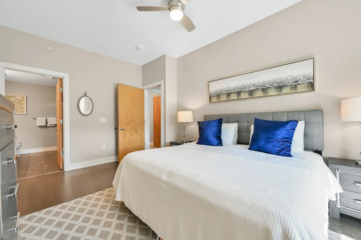 A serene king-sized bedroom featuring a plush bed adorned with decorative pillows, a nightstand with a lamp, and tasteful artwork on the wall. The room is softly lit with natural light, creating a calm and restful atmosphere.