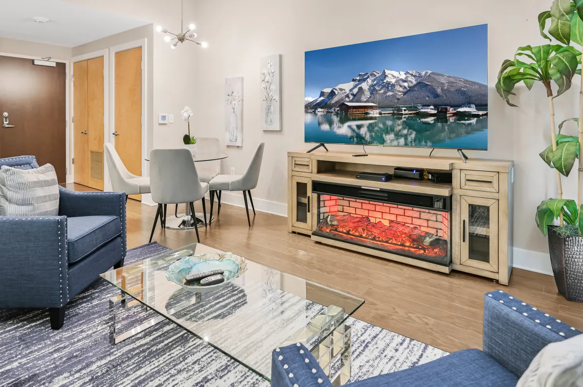 A modern living room featuring a large television displaying a scenic mountain view, a decorative fireplace, and a glass coffee table. The space is bright, inviting, and set up for relaxation or casual gatherings, suggesting a comfortable return after city adventures.