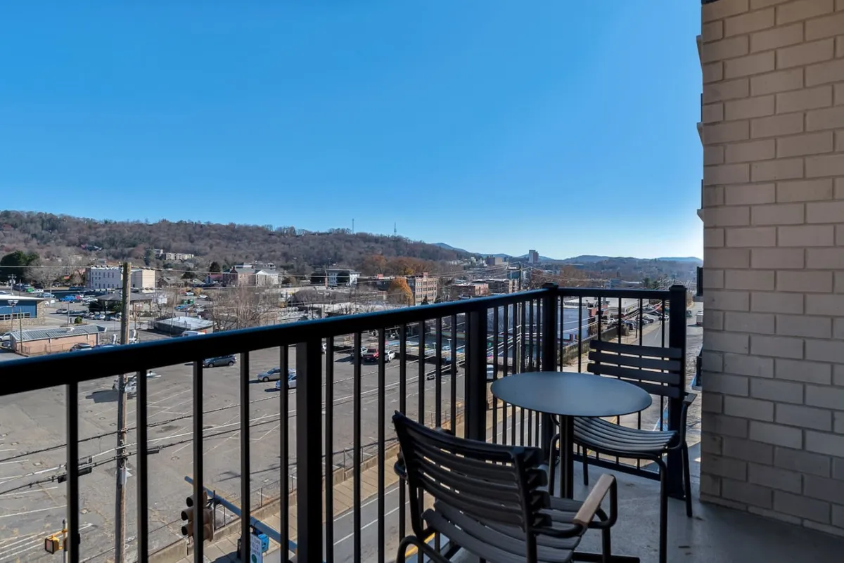 This exterior-focused image shows the private balcony with a small table and chairs, looking out over the city with distant hills on the horizon. The scene evokes tranquility and connection to Asheville's urban and natural environment, perfectly matching the description of enjoying morning coffee or an evening drink while taking in the view.