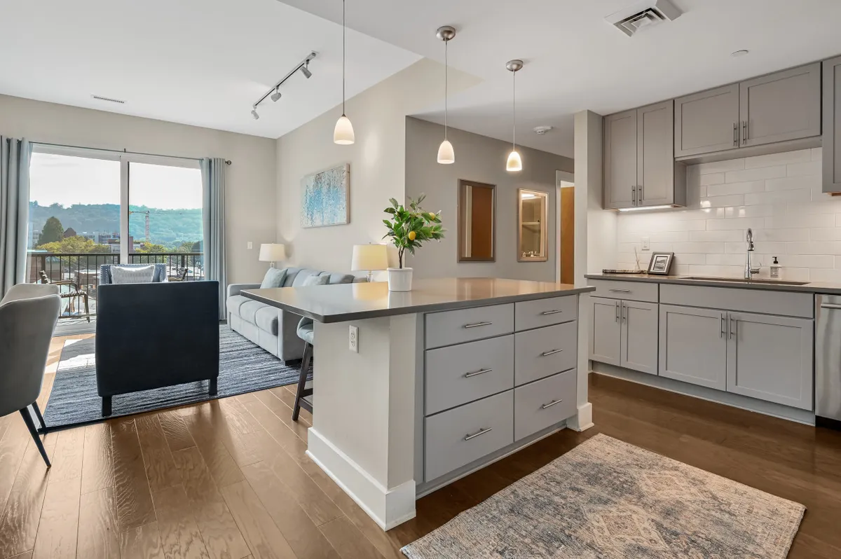 This image captures the open-concept living area, seamlessly blending the kitchen island, a comfortable seating area with a sofa, and a clear view out to the private balcony. Modern furnishings and decorative art lend a stylish, urban vibe, while the sunset visible through the balcony doors suggests a relaxing, welcoming environment perfect for unwinding or entertaining.