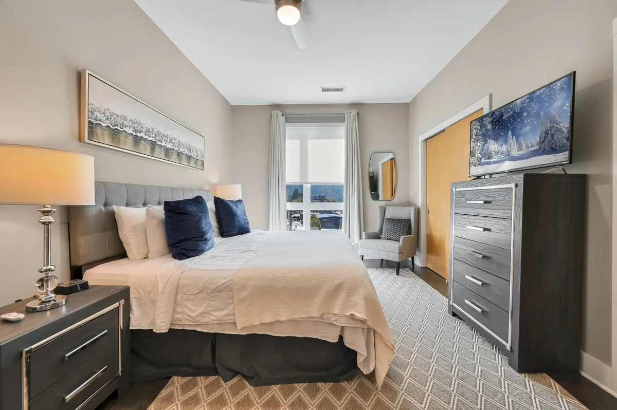 Another elegant king bedroom with white bedding and dark blue accent pillows, a nightstand with a lamp, a dresser with television, and a window draped with curtains. The room exudes comfort and privacy.