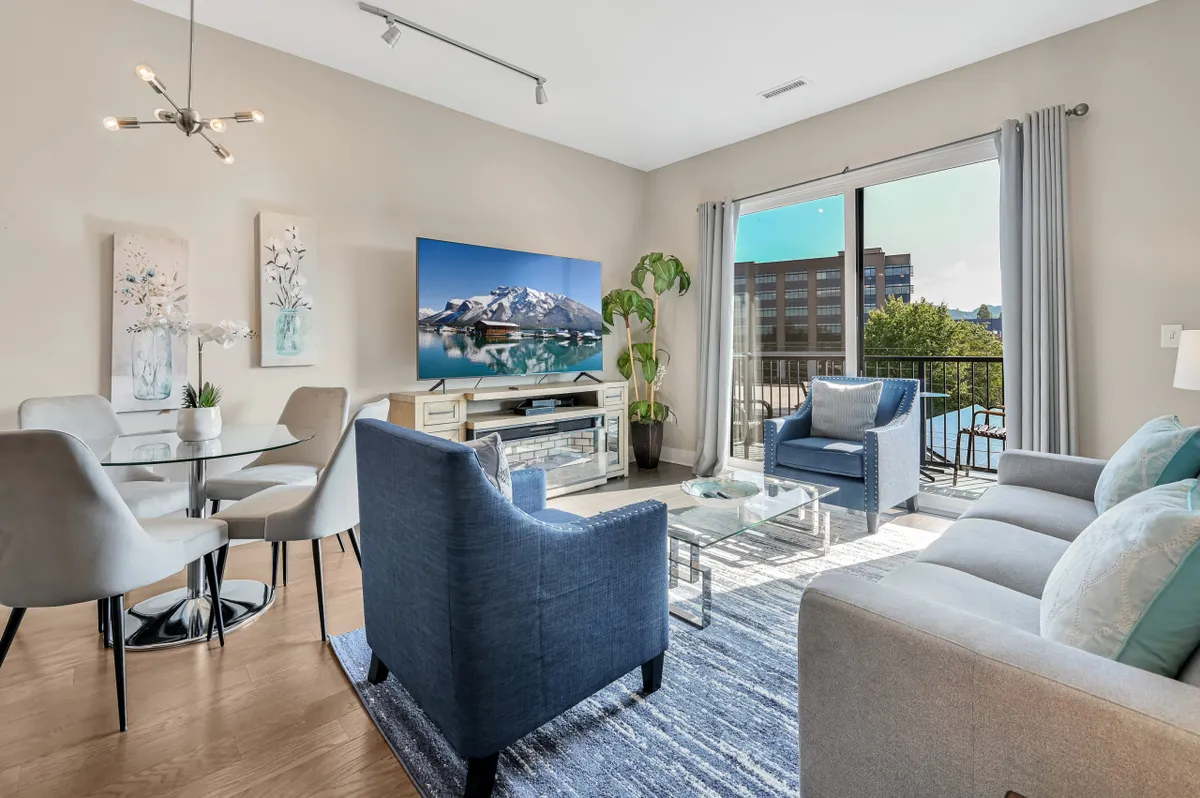 The image showcases a bright, modern living area with a plush sofa, inviting armchairs, a stylish dining table, and a large window that fills the space with natural light. The setup is perfect for relaxing, watching TV, sharing meals, or gathering with friends and family, all within a cozy yet contemporary ambiance.